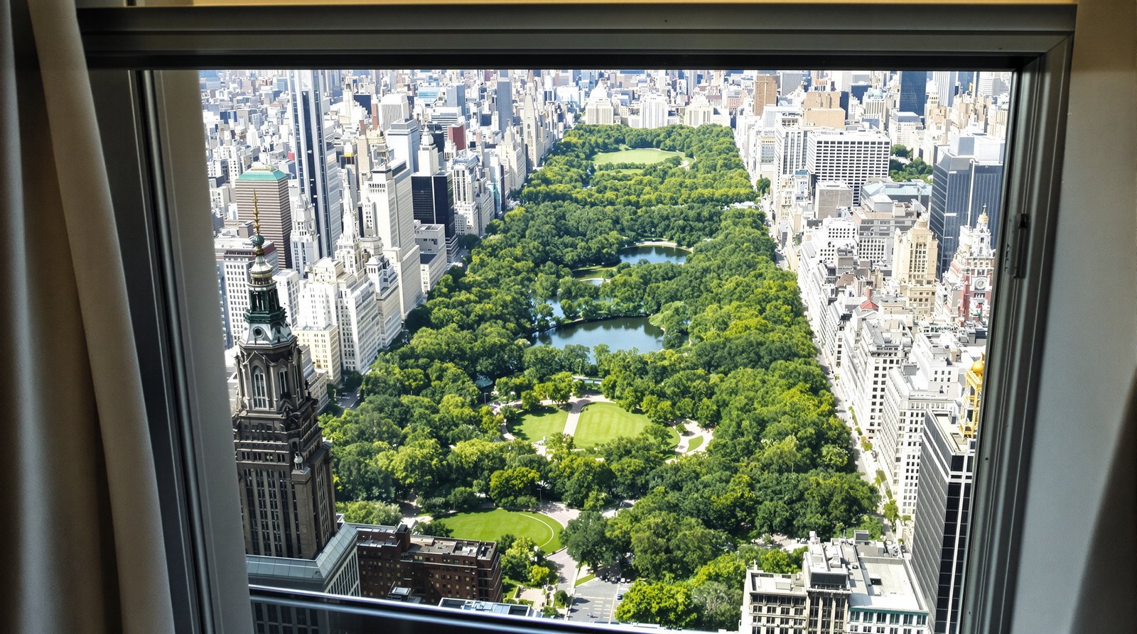 A stunning, ultra-realistic view of Central Park from a high-floor hotel room window, with the park's greenery contrasting with the surrounding city buildings under bright, natural daylight