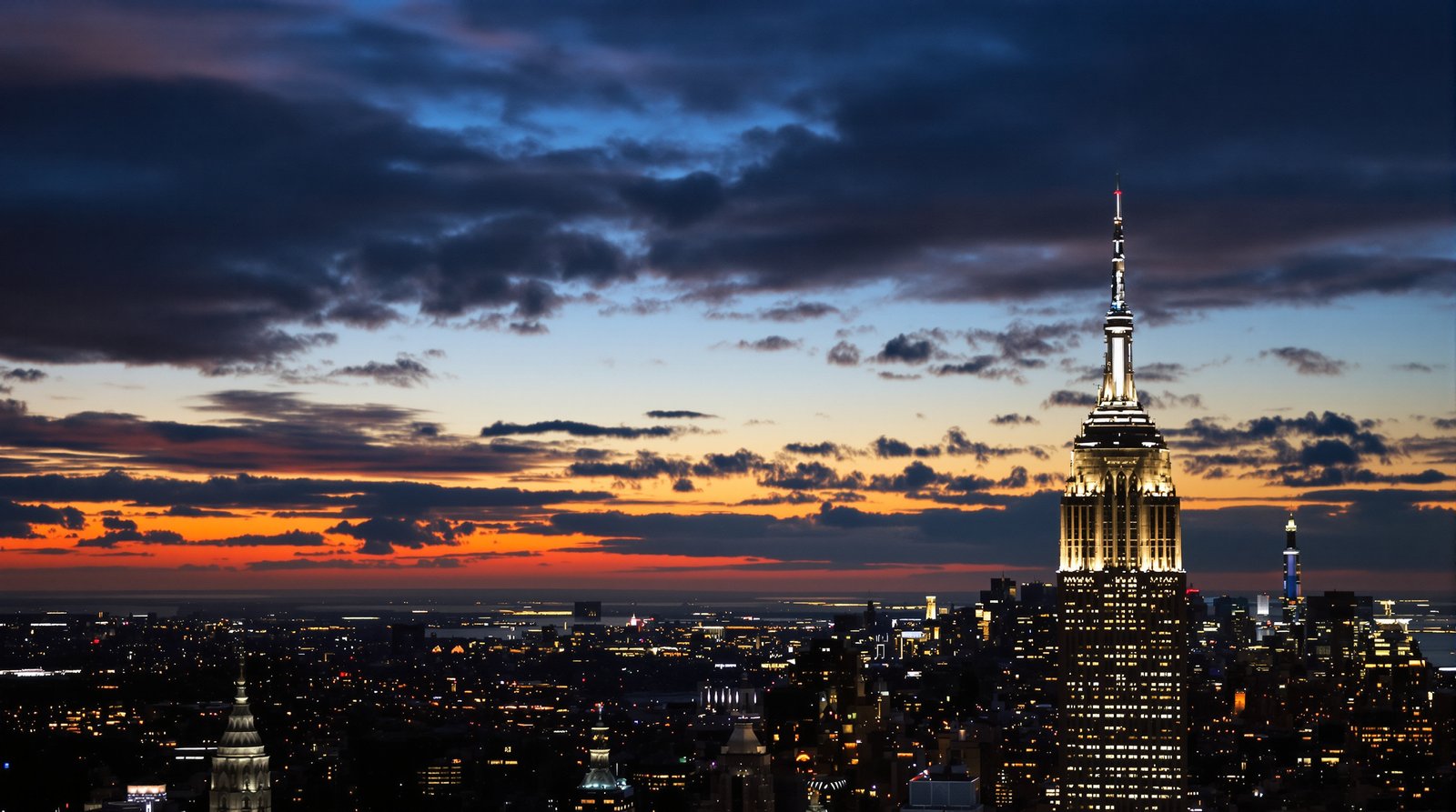 The iconic spire of the Empire State Building piercing the clouds at sunset, with the sprawling city lights beginning to twinkle below