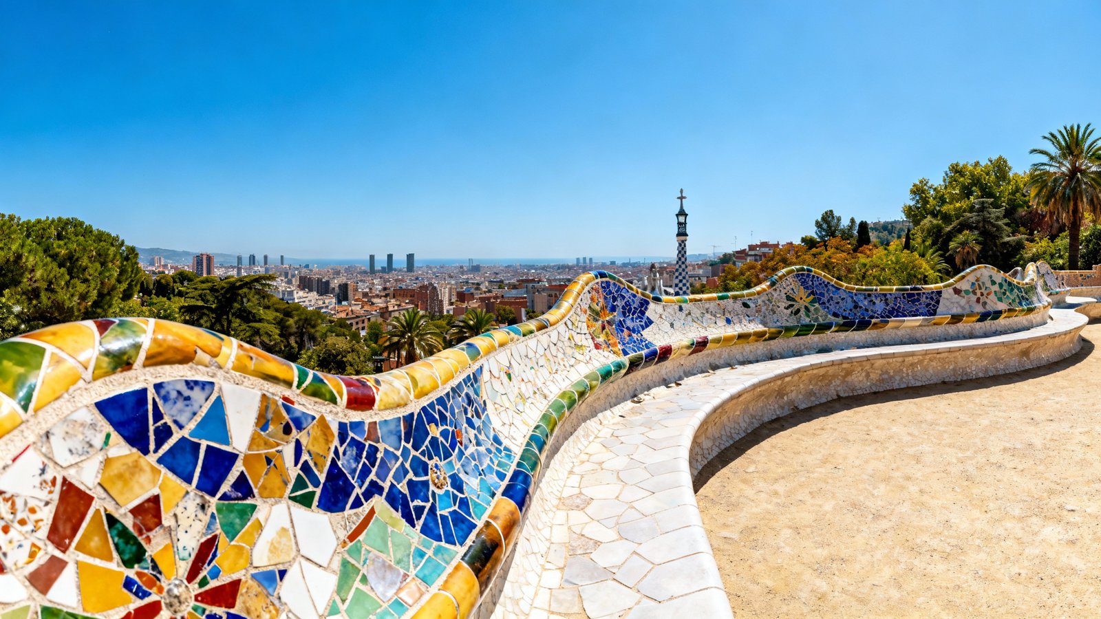 A wide-angle shot of the mosaic-covered serpentine bench in Park Güell, with the Barcelona skyline in the background under a clear blue sky